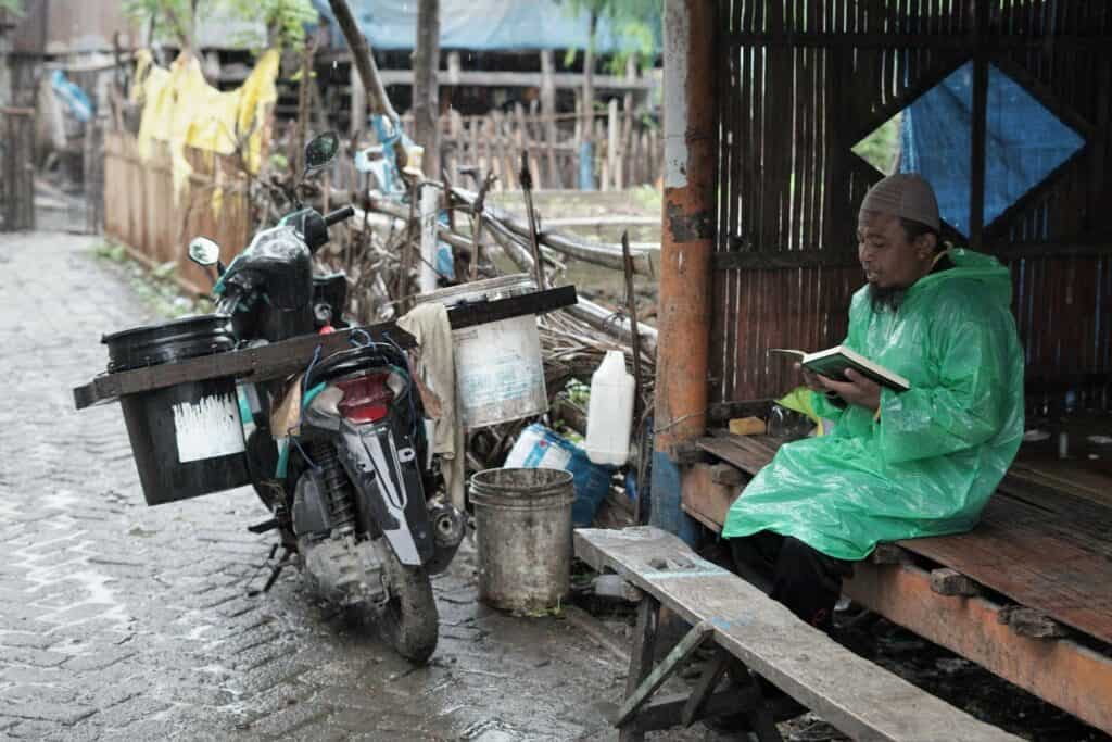 Busron Daeng Kawang menyempatkan diri membaca Al-Qur’an di sela aktivitasnya berjualan ikan keliling dari kampung ke kampung di wilayah Kelurahan Biringkassi, Kecamatan Binamu, Jeneponto, menggunakan sepeda motornya sejak selepas solat subuh, jam 05.00-08.00 pagi.